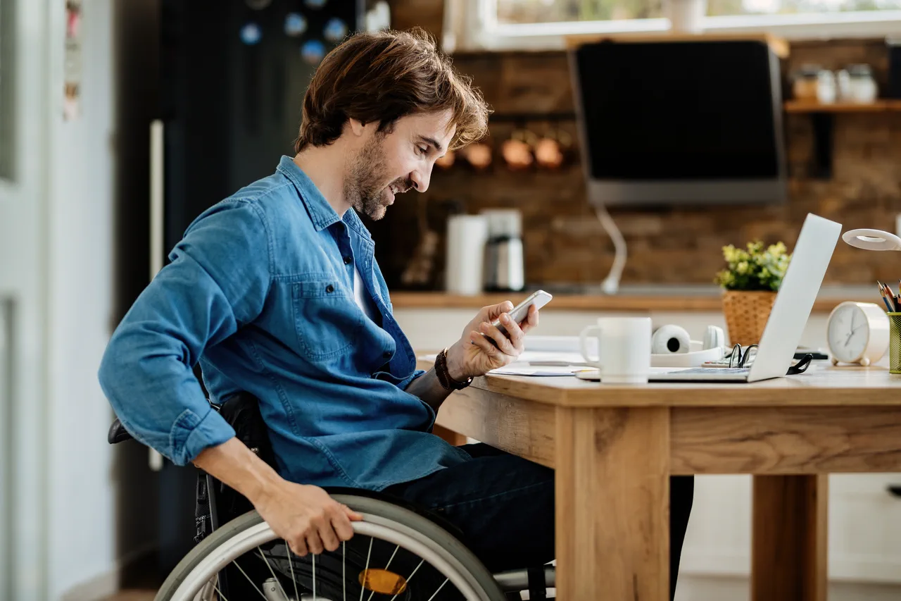 A man in a wheelchair reading a message on his phone while working on a laptop at home.
