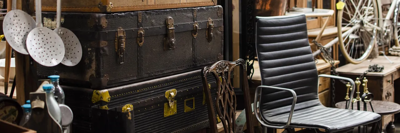 Old leather trunks and suitcases stacked in an antiques market, beside a vintage bicycle and an office chair.