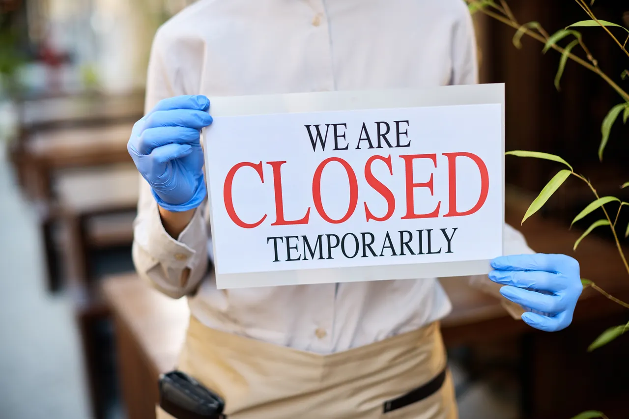 A person in blue gloves holding a sign that says We Are Closed Temporarily.