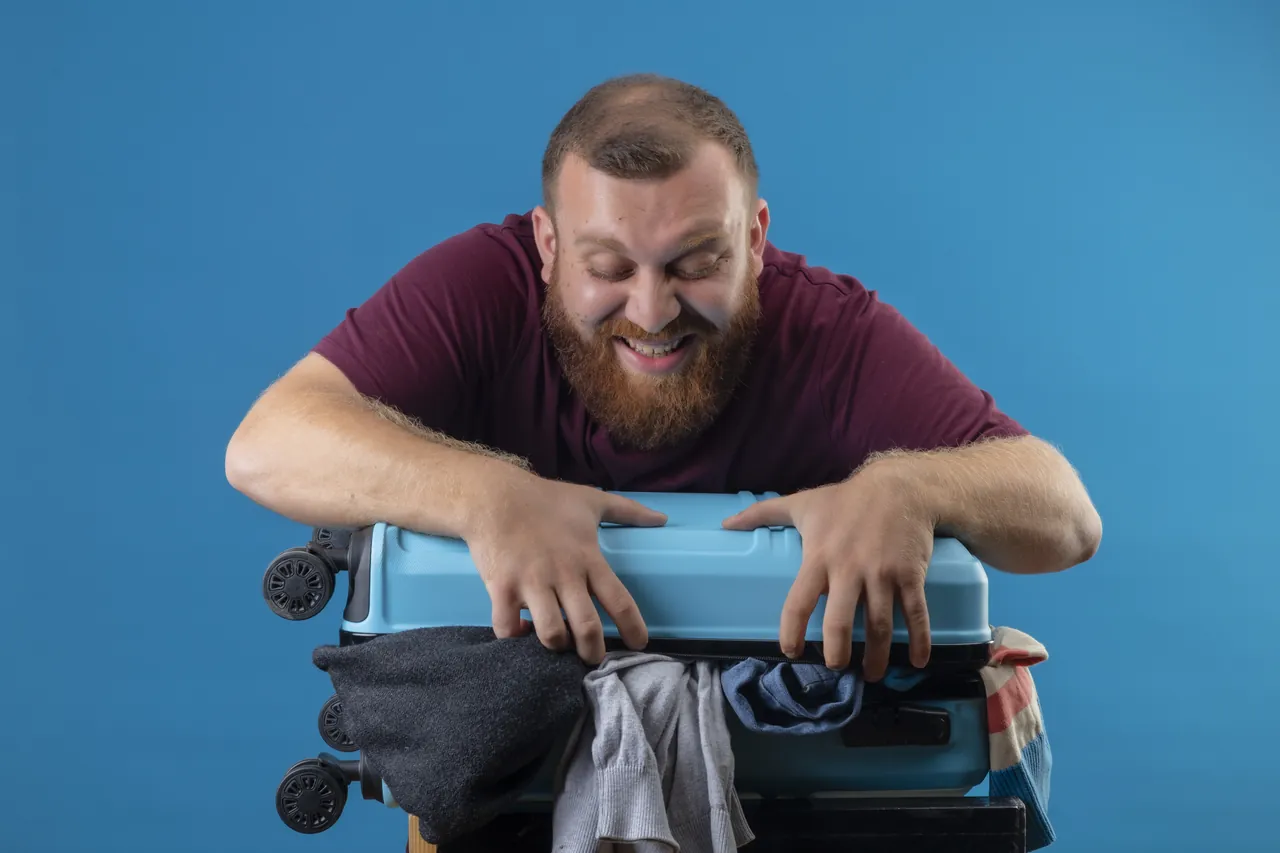 A man leaning on an overstuffed suitcase, trying to close it.